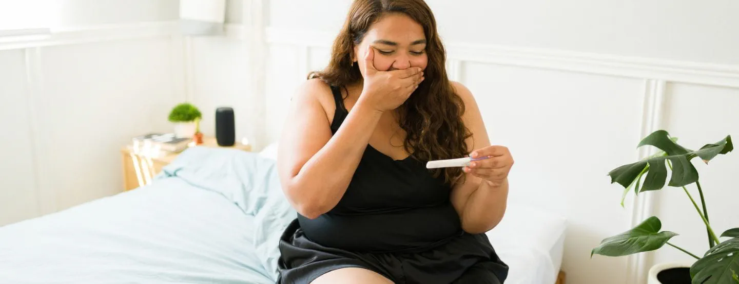 Woman sitting on bed looking at pregnancy test with surprise and happiness. 