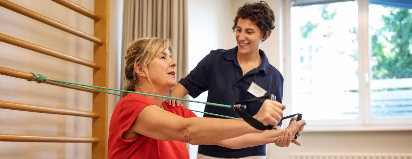 Woman using therapy bands to exercise arms while a healthcare professional guides her.