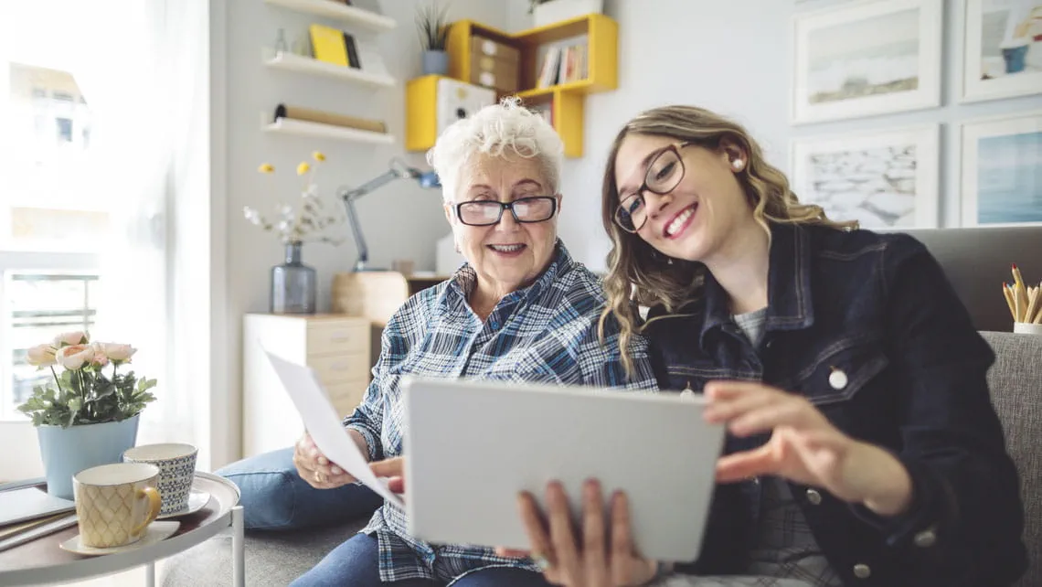 a woman and her adult daughter view a tablet together on a video chat
