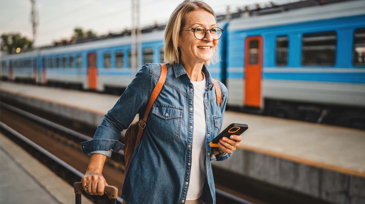 a woman stands on a train platform with a suitcase and cell phone