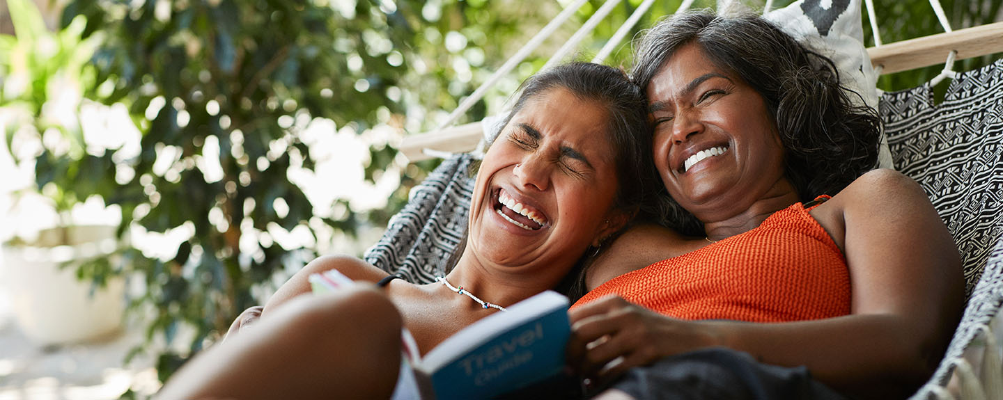 a mother and her adult daughter laugh together outdoors on a hammock