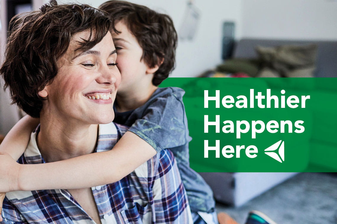 A young boy embraces his mother. Overlay text reads "Healthier Happens Here."