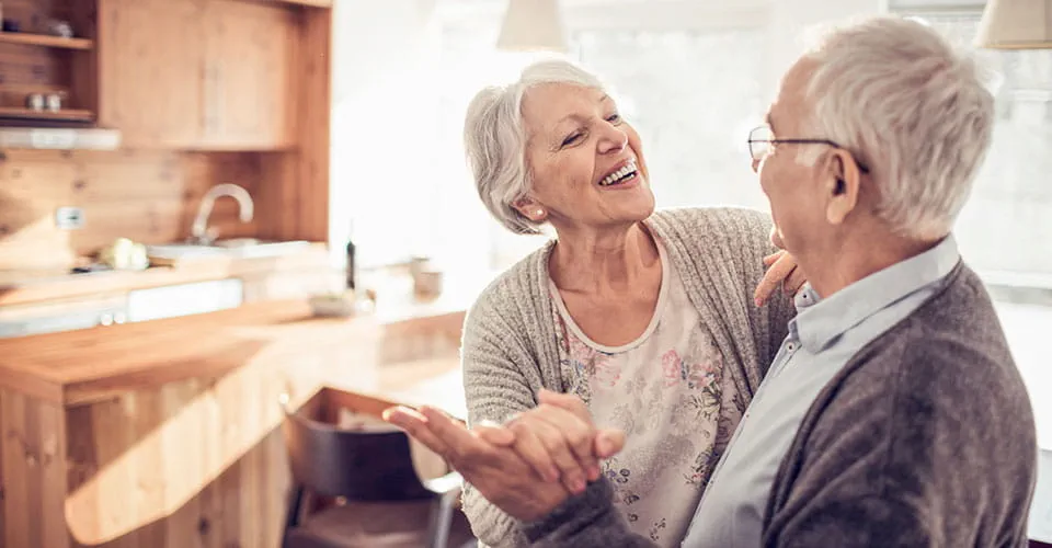 A senior couple hold hands and laugh while dancing in their home